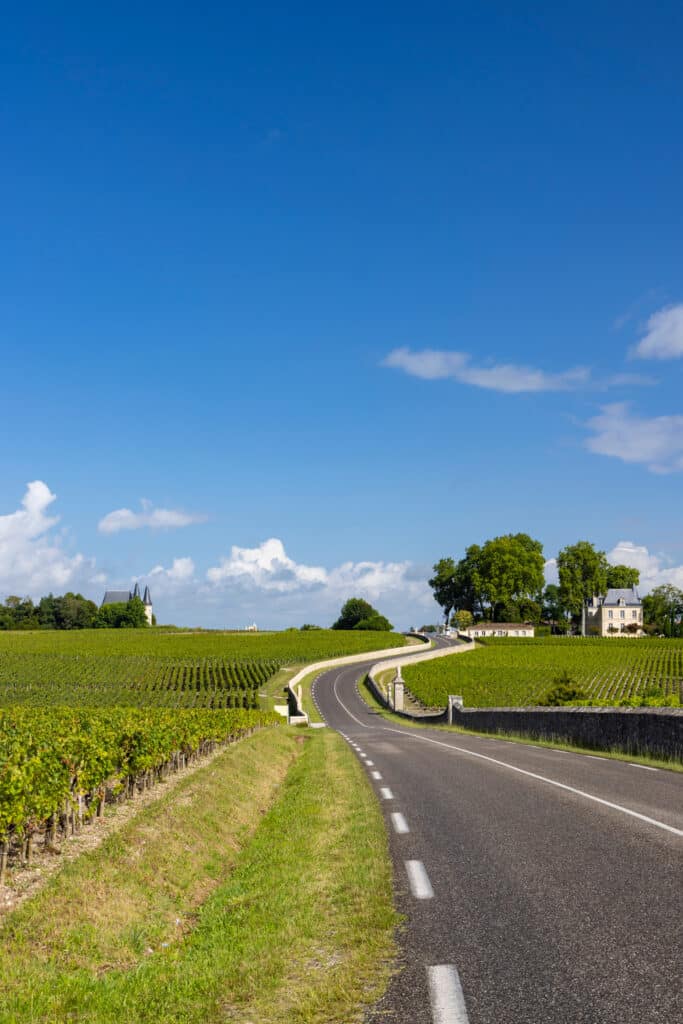 Vignobles de Bordeaux : Route vers le Château Route sinueuse traversant des vignobles luxuriants vers un élégant château bordelais sous un ciel bleu et nuageux.