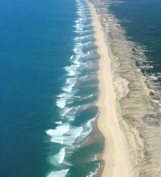 Plage Atlantique Vagues et Dunes Côtières Vue aérienne d'une longue plage de sable bordée par des vagues bleues déferlantes et des dunes boisées en arrière-plan.